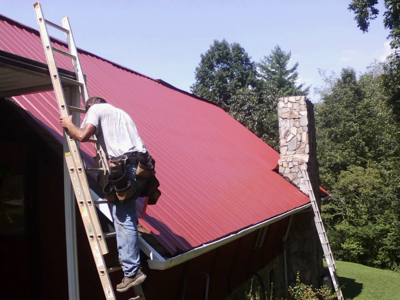 Roofer on a ladder beside a red metal roof and stone chimney — Soffit & Fascia Repair by Roofing Brothers in Joelton